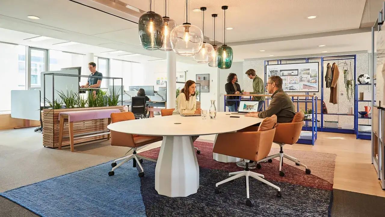 Un espace convivialit&eacute; entreprise moderne avec une grande table ovale, des fauteuils confortables et des plantes vertes. Des collaborateurs discutent autour de la table.