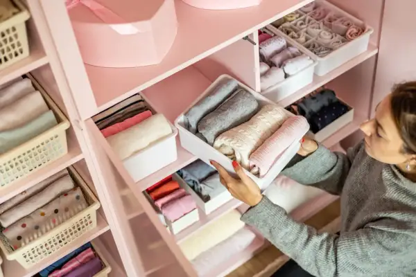 Une femme range des vêtements pliés dans des boîtes dans une armoire rose. Une excellente idée rangement pour organiser ses affaires.