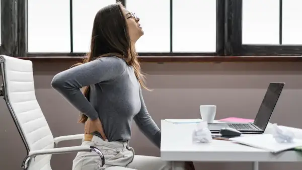 Femme se tenant le dos, visiblement inconfortable. Elle a du mal à bien s'asseoir à son bureau.
