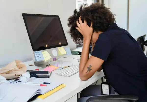 Une femme aux cheveux bouclés est stressée au travail, les mains dans les cheveux, devant un bureau encombré. Des post-it sur l'écran indiquent "étape 4 finaliser les assets".