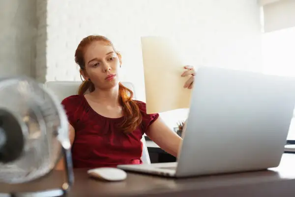 Femme rousse transpirant au bureau, ventilateur en marche, pour lutter contre la température maximale.
