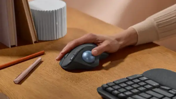 Maintenant, quelle souris ergonomique choisir ? Une main contrôle une souris grise avec une boule bleue sur un bureau en bois.